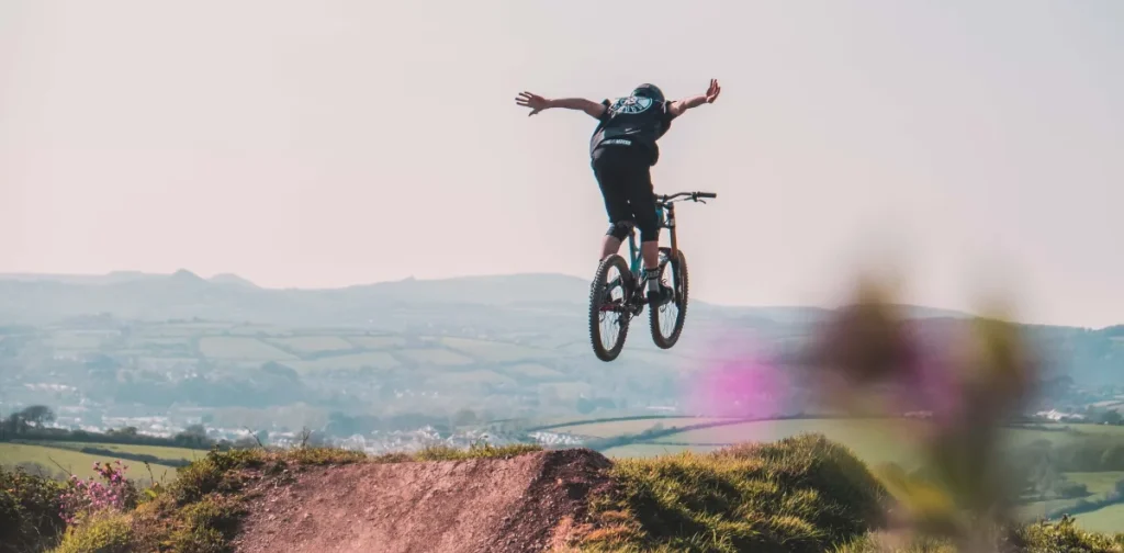 A mountain biker performing a jump over a dirt ramp with a scenic landscape in the background.