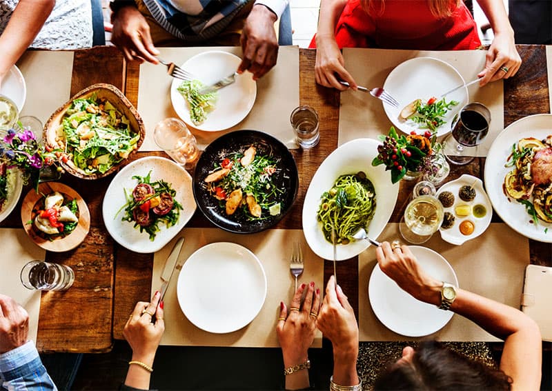 People sitting around a wooden table sharing a colorful meal with salads, pasta, grilled food, and drinks. Hands are reaching for food, creating a lively, communal dining atmosphere.