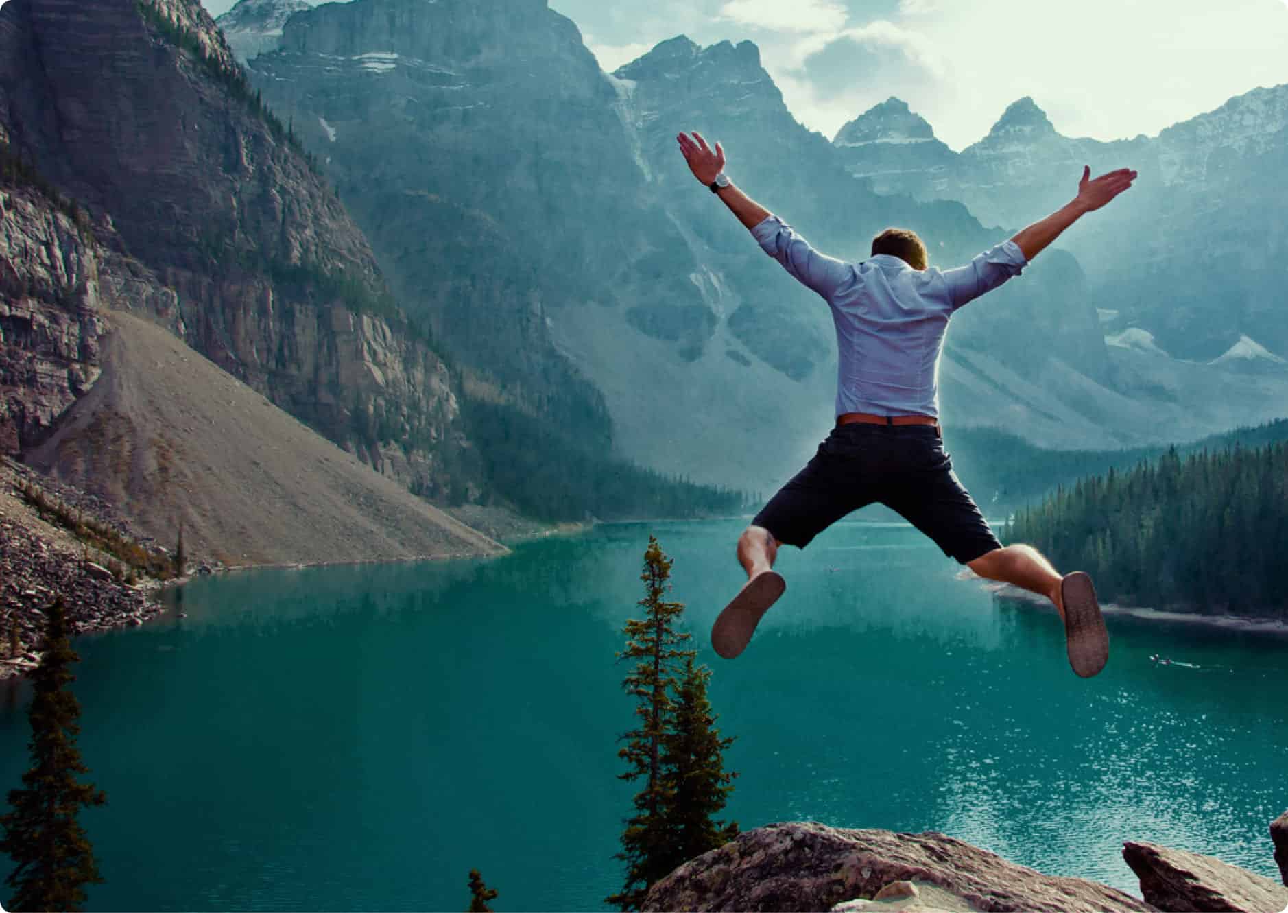 A person wearing a blue shirt and shorts jumps joyfully off a rock, overlooking a turquoise lake surrounded by pine trees and rugged mountain peaks under a cloudy sky.