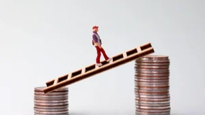A miniature figure walking on a wooden plank balanced between stacks of coins.