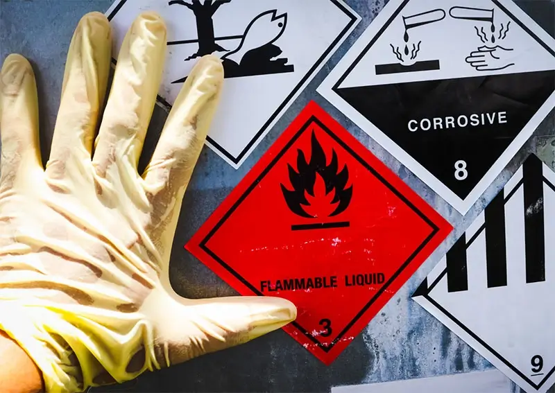 A gloved hand is shown next to warning signs for hazardous materials, including a red flammable liquid sign, a corrosive symbol, and other caution labels.