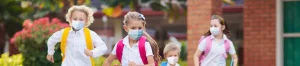 group of children wearing masks running towards school with backpacks