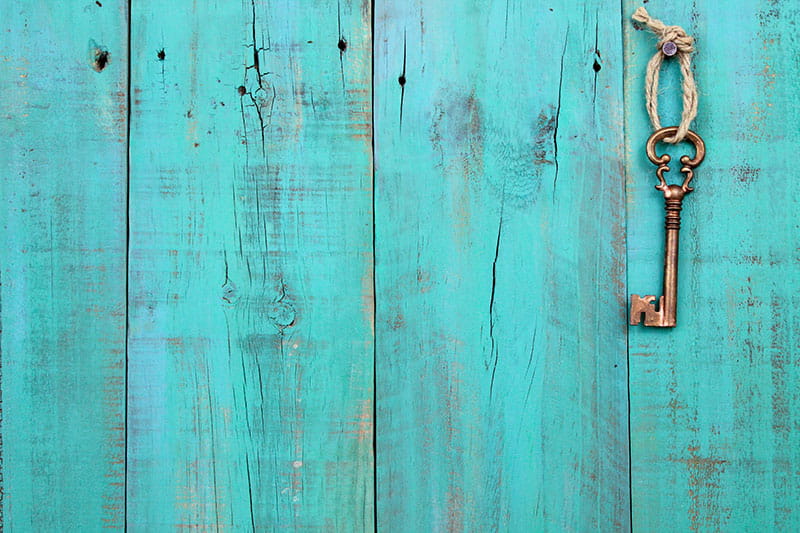 A vintage brass key tied with rope hangs from a nail on a weathered turquoise wooden plank wall.