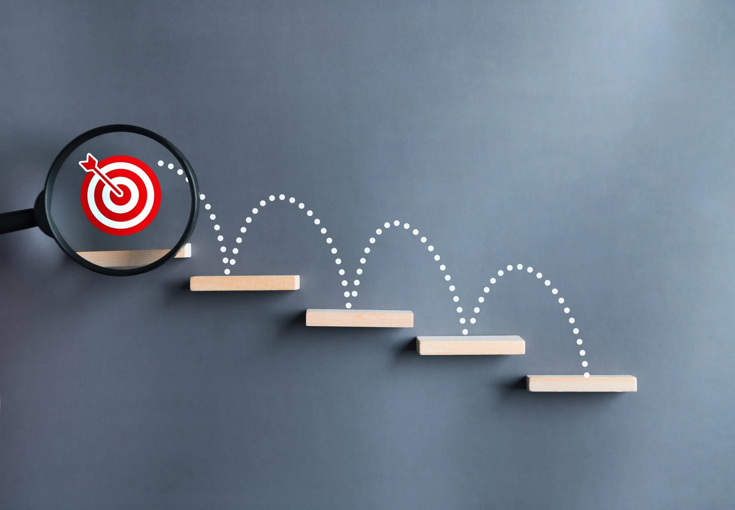 A magnifying glass focuses on a red target symbol at the top of a staircase made of wooden steps, with a dotted line illustrating a path upward over each step.