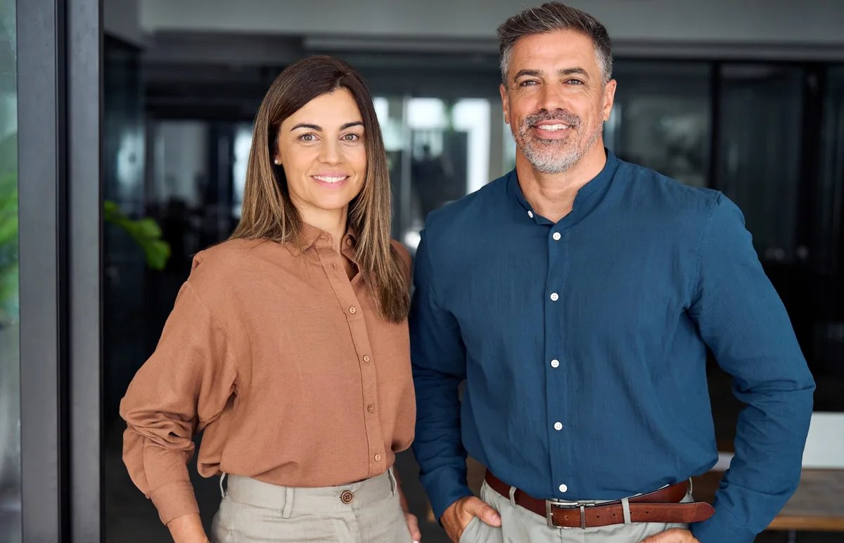 A woman and a man stand side by side indoors, both smiling at the camera. The woman wears a brown shirt and beige pants, while the man wears a blue shirt and gray pants. They appear to be in an office setting.