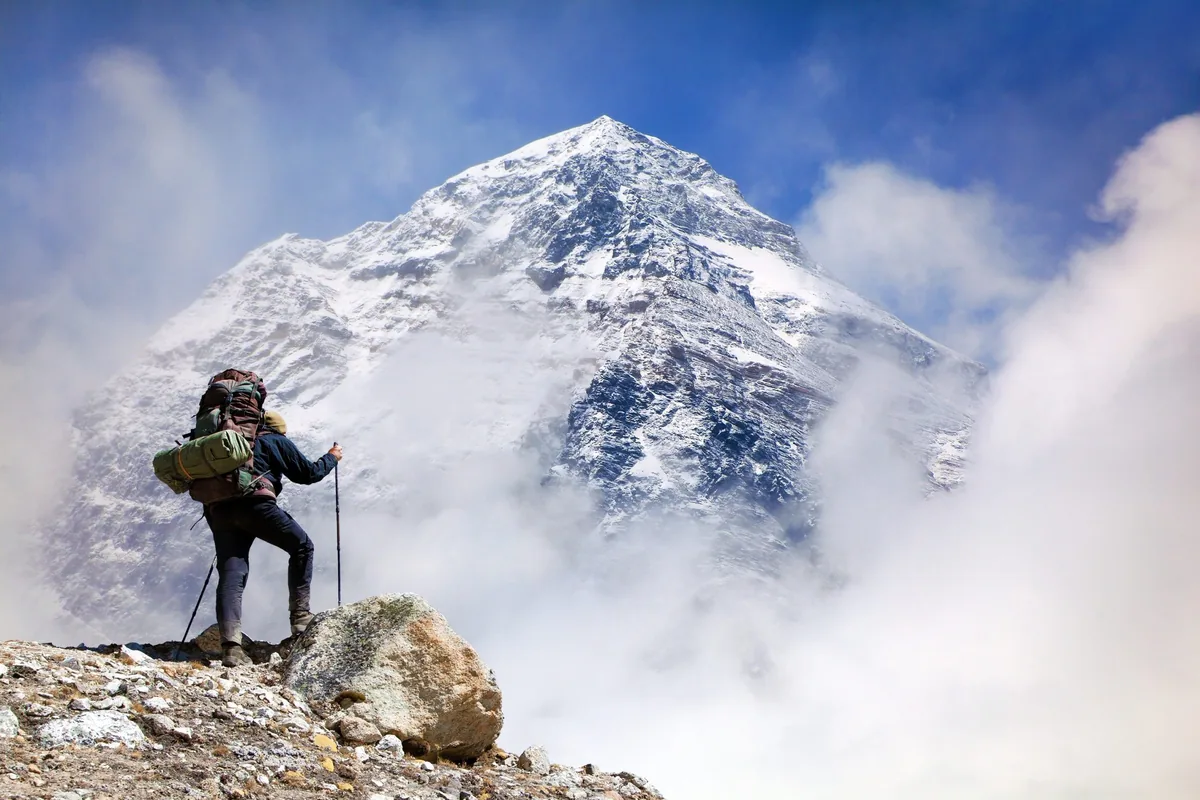 A hiker with a large backpack and trekking poles standing on a rocky ridge, looking toward a massive, snow-covered mountain peak partially obscured by clouds.