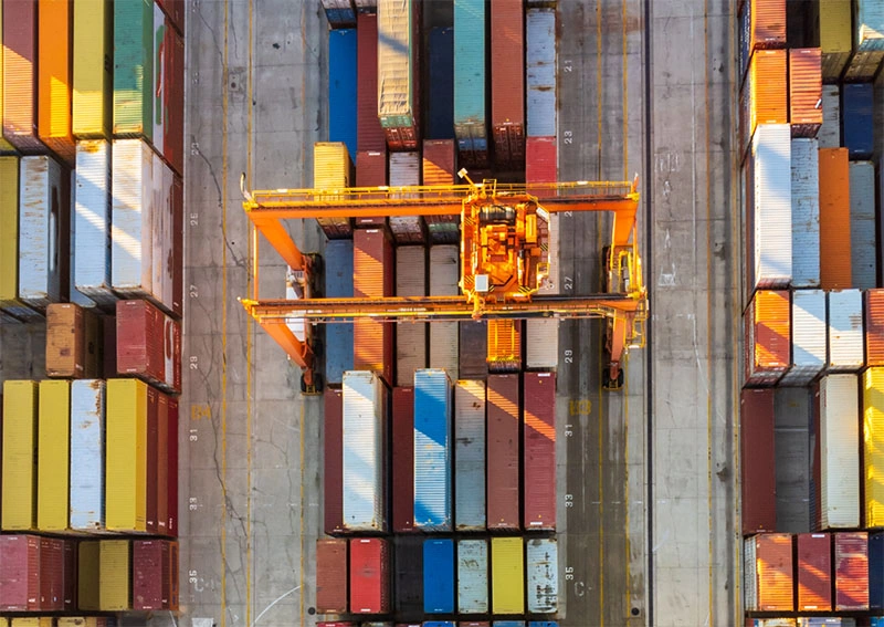 Aerial view of a shipping container yard with a large yellow crane lifting containers among neatly arranged rows of multicolored containers on concrete ground.
