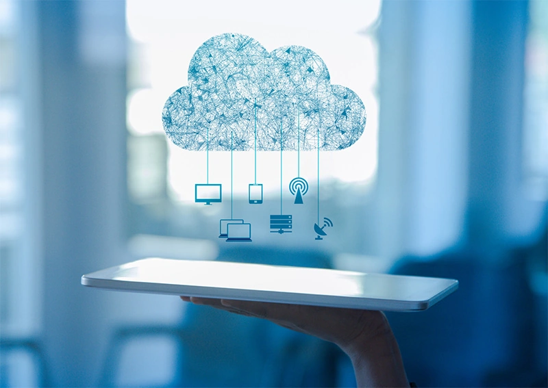A hand holds a tablet, above which a digital cloud icon appears with connected symbols for devices and communication, representing cloud computing and data connectivity. The background is blurred and blue-toned.
